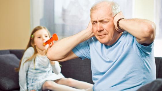 Ein älterer Herr sitzt auf einem Sofa und presst die Hände auf seine Ohren, während ein Mädchen hinter ihm in eine Spielzeugtröte bläst.