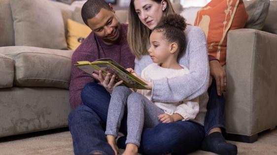 Junge Familie sitzt zusammen und die Eltern lesen dem Kind aus einem Buch vor.