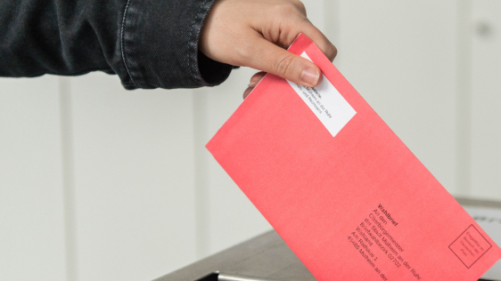 Eine Hand wirft den versiegelten roten Wahlumschlag mit dem Stimmzettel in den Schlitz der Wahlurne.