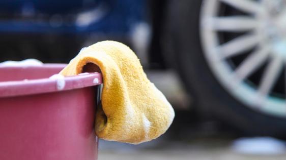 Im Vordergrund ist ein mit Putzmittel befüllter Eimer abgebildet, über dessen Rand ein Schwamm hängt. Im Hintergrund sieht man ein Fahrzeug.