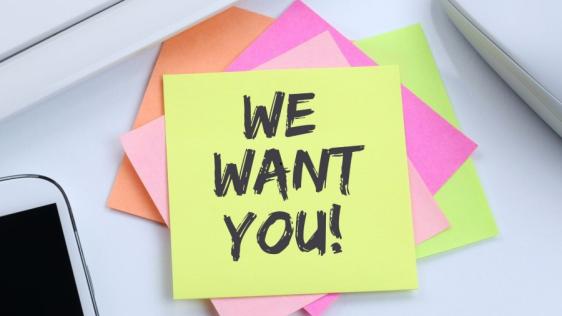Mehrere bunte Klebezettel liegen übereinander auf einem Schreibtisch. Auf dem oberen Zettel steht We want you!