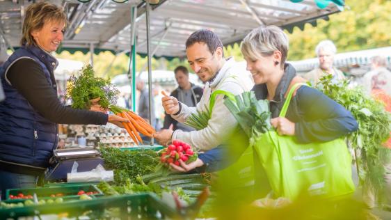 Szene an einem Marktstand. Ein Mann und eine Frau kaufen bei einer Händlerin Radieschen und Möhren. 