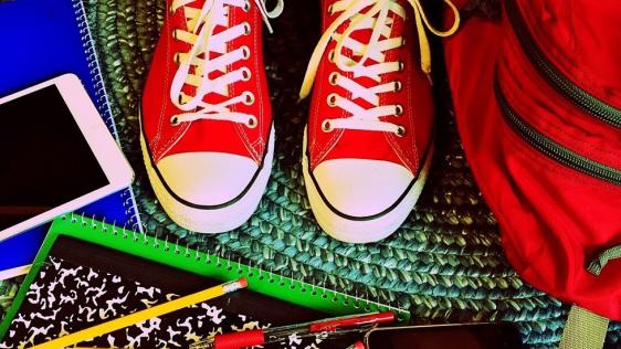 Rote Turnschuhe auf einem Teppich, rundherum liegt Schulmaterial wie Stifte, Hefte, roter Schulranzen und mehr.