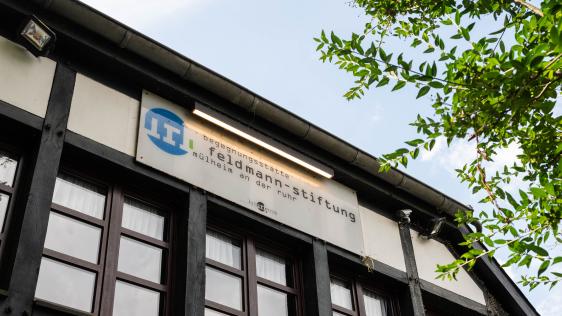 Schild mit Text Feldmann-Stiftung an Fachwerkhaus mit Fenstern und grünem Ast im Vordergrund