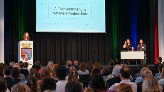 Auf der Bühne begrüßt Julia Brunner zusammen mit ihrem Team (Lara Vercoulen und Heiko Möller) die Teilnehmenden der Auftaktveranstaltung Netzwerk Kinderschutz 