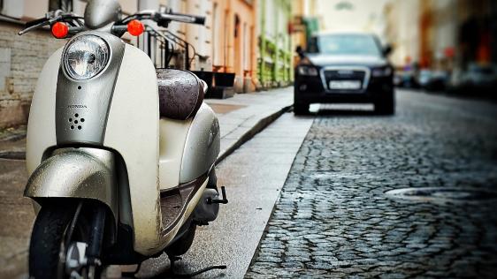 Das Foto zeigt einen Motoroller und ein Auto geparkt in einer Straße mit Kopfsteinpflaster umringt von Altbauten.