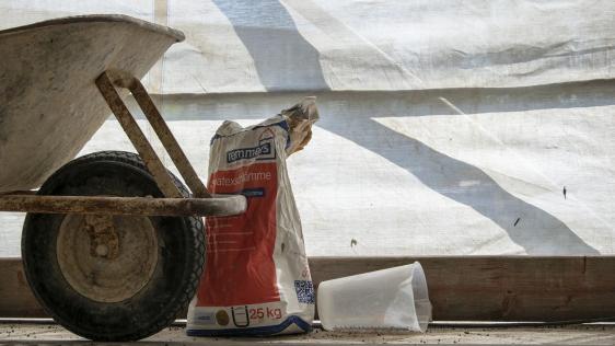 Bildausschnitt einer Baustelle mit einer Schubkarre, einem Sack Beton und anderem Baumaterial.