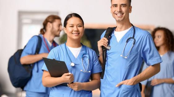 Eine Medizinstudentin und ein Medizinstudent stehend lächelnd in der Universität. Im Hintergrund befinden sich zwei weitere Student*innen.