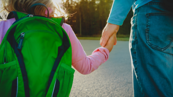 Bildausschnitt mit einem Elternteil, der sein kleines Kind mit grünem Rucksack an der Hand hält und aus dem Bild laufen. Tagespflegedienst, Kita, Kindergarten, Kinderbetreuung, Pflegevater, Pflegemutter, Kindertagespflege.