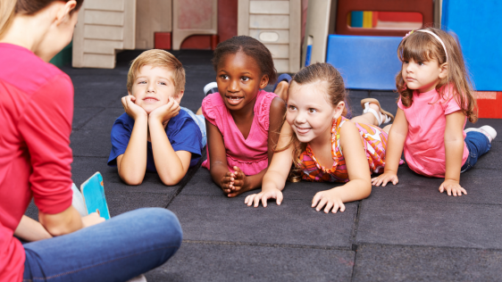 Tagespflegeperson sitzt vor vier Kindern, sie liest vor und die Kinder hören zu.