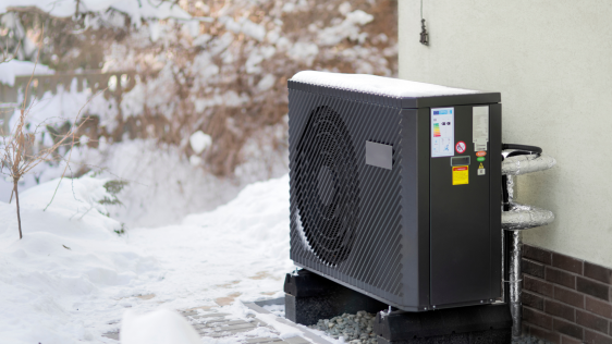 Dunkelgraue Wärmepumpe an einer Hauswand mit Blick in einen verschneiten Garten.