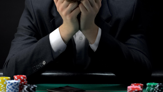 Mann sitzt am Pokertisch, vor sich eine leere Brieftasche, und vergräbt den Kopf verzweifelt in den Händen