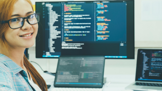 Eine junge Frau sitzt vor Bildschirmen, die Programmierprozesse zeigen und schaut freundlich 