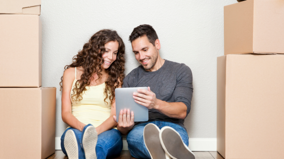 Ein Mann und eine Frau sitzen auf dem Boden zwischen Umzugskartons, schauen auf ein Tablet und unterhalten sich gut gelaunt über das, was sie sehen