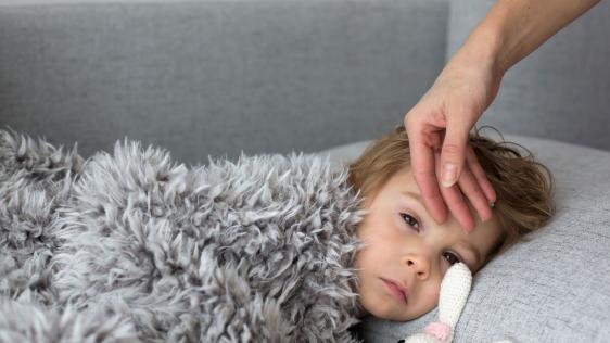 Kind liegt krank auf der Couch. Daneben liegt ein Kuscheltier