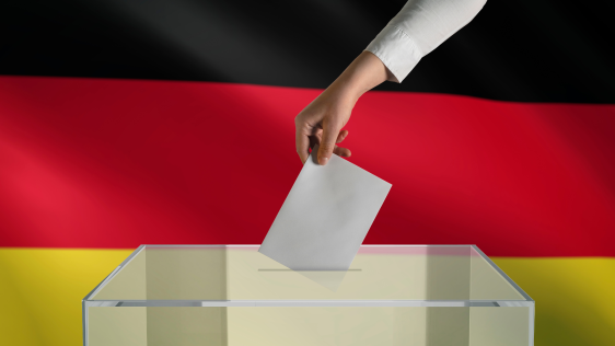 eine Person wirft einen Wahlbrief in eine Urne. Im Hintergrund eine Deutschlandflagge