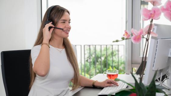 Frau mit Headset sitzt am Schreibtisch im Kommunikations Center. Vor ihr steht ein Computer und eine Tasse Tee. Im Hintergrund sind Pflanzen und Fenster zu sehen.