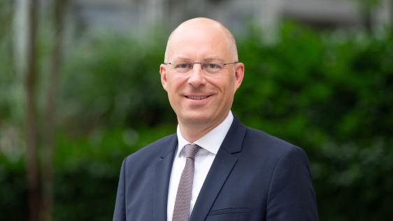 Felix Blasch trägt einen dunklen Anzug und eine Krawatte, steht vor einer grünen, unscharfen Hintergrundlandschaft.