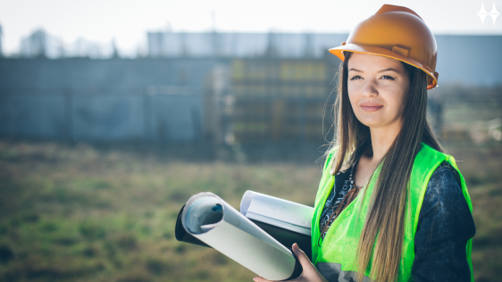 Das Bild zeigt eine junge Frau, die mit Helm und Warnweste in einer Baulandschaft steht. In ihren Händen hält sie einen aufgerollten Plan.