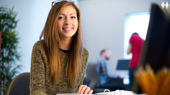 Das Bild zeigt eine junge Frau sitzend an einem Tisch. Auf dem Tisch vor ihr liegt ein aufgeschlagener Aktenordner. Sie lächelt in die Kamera.