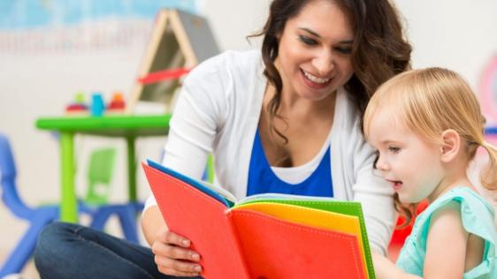 Eine Frau sitzt mit einem kleinem Mädchen in einem Spielzimmer und schaut sich ein Kinderbuch an.