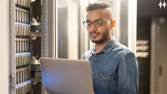 Das Bild zeigt einen jungen Mann stehend vor einem Schaltschrank. Er hält ein Laptop in den Händen und blickt konzentriert darauf.