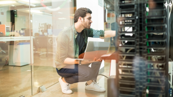 Das Bild zeigt einen jungen Mann hockend vor einem Schaltschrank. Er hält ein Laptop in den Händen und blickt konzentriert auf den Schaltschrank..