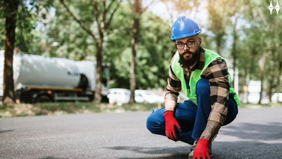 Das Bild zeigt einen jungen Mann, der in Warnweste mit Helm und Handschuhen auf einer asphaltierten Straße hockt. er schaut in die Kamera