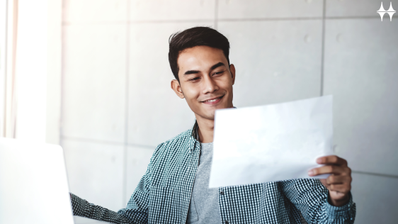 Das Bild zeigt einen jungen Mann sitzend an einem Tisch. In seiner linken Hand hält er ein weißes Blatt Papier. Auf dem Tisch steht ein aufgeklapptes Laptop. 
