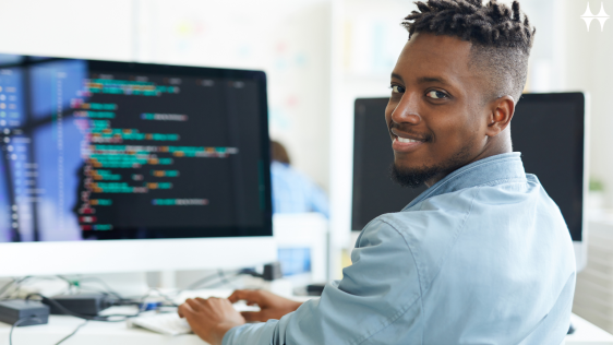 Das Bild zeigt einen jungen Mann sitzend an einem Tisch. Er dreht sich in die Kamera und lächelt. Auf dem Tisch steht ein Monitor und eine Tastatur. Der Monitor zeigt einen bunten Programmtext.