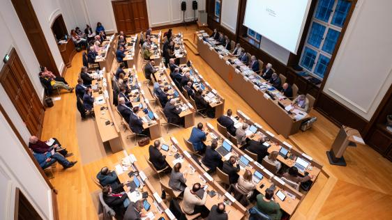 Rat der Stadt tagt im Ratssaal mit mehreren Reihen von Tischen, an denen zahlreiche Personen sitzen und diskutieren. Stadt Mülheim an der Ruhr