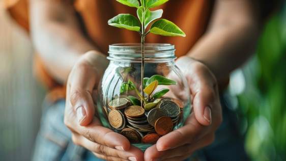 2 Hände halten einen Glasbehälter in dem sich Münzen befinden und ein junger Pflanzentrieb herauswächst