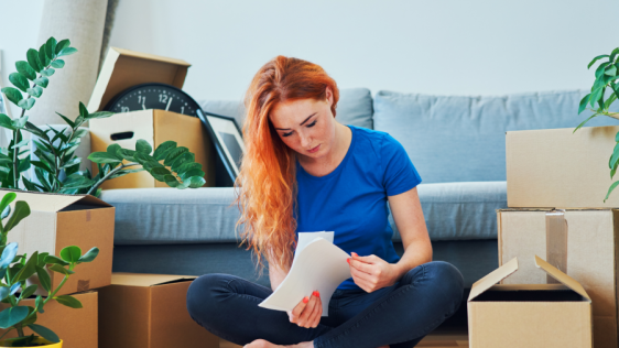 Junge Frau sitzt vor Umzugskartons und Couch, hält Unterlagen in der Hand. Kosten der Unterkunft
