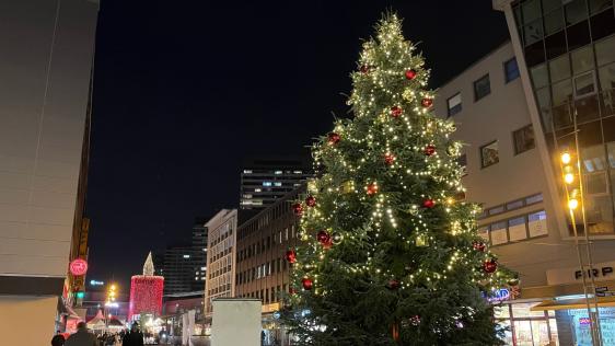 Weihnachtsbaumdekoriert mit Kugeln und Lichterketten und im Hintergrund ist ein Weihnachtsmarkt