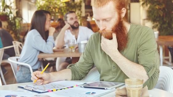 Junger rothaariger Mann mit Bart im Cafè der am Tisch über einem Tablet und Geschäftszahlen grübelt