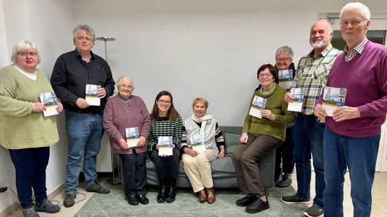 Die Mitglieder der Arbeitsgruppe Dümpten aus dem Netzwerk der Generationen haben sich für ein Gruppenbild aufgestellt. In der Mitte sitzen einige Personen auf einem Sofa, links und rechts daneben stehen weitere Personen und halten den neuen Wegweiser in die Kamera.