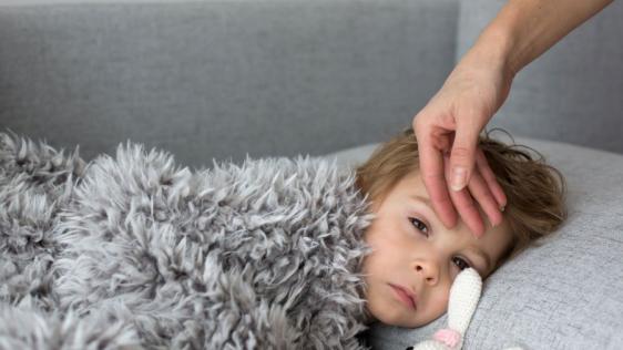 Ein krankes Kind liegt mit seinem Stoffhasen und einer Decke auf einer Couch. Die Mutter fühlt mit ihrer Hand, ob es Fieber hat.