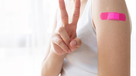 Eine junge Frau mit einem weißen Shirt wurde geimpft. Auf Ihrem Oberarm befindet sich ein pinkes Pflaster.