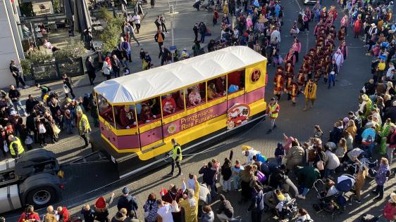 Blick aus dem Rathaus auf den Rosenmontagszug in der Schollenstraße. Der Mottowagen der Prinzengarde Rote Funken sieht aus wie eine alte Straßenbahn. Die Karnevalist*innen werfen Kamelle, die die Kinder und Erwachsenen am Straßenrand aufsammeln.