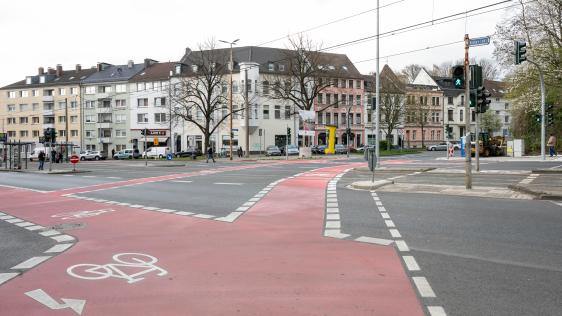 Breiter roter Radweg mit Fahrradpiktogrammen an einer Kreuzung vor mehrstöckigen Wohngebäuden und Fußgängern
