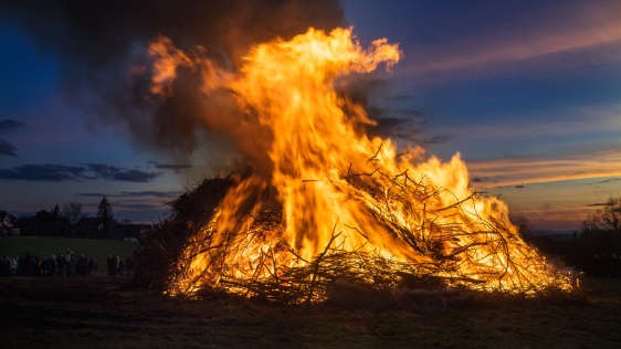 Ein riesiges Osterfeuer brennt in der Dämmerung auf einer Wiese. Rauch und Flammen steigen in die Luft. Links im Hintergrund sind die Umrisse von Menschen zu erkennen.
