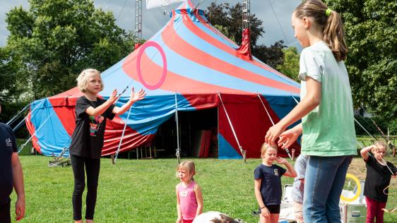 Kinder balancieren auf Bällen vor einem Zirkuszelt.