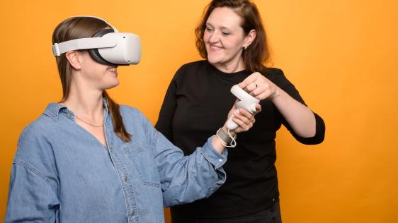 Zwei Personen sind zu sehen. Eine davon trägt eine VR-Brille und interagiert mit einer virtuellen Umgebung, die andere steht daneben.