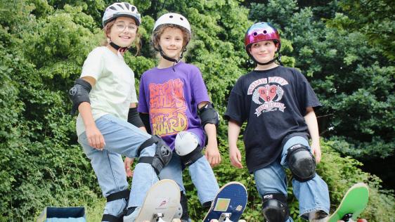 Drei Mädchen mit Helm und Schutzausrüstung stehen mit Skateboards im Grünen und posieren gemeinsam für ein Foto.