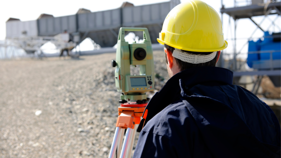Auf dem Bild sieht man einen Mann mit Helm von hinten. Vor ihm steht ein Vermessungsgerät. Er steht auf einer Baustelle.