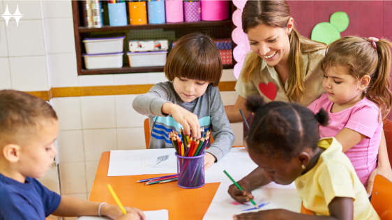 Auf dem Bild sitzen vier Kinder an einem Tisch und malen. Eine junge Frau kniet daneben und lächelt.
