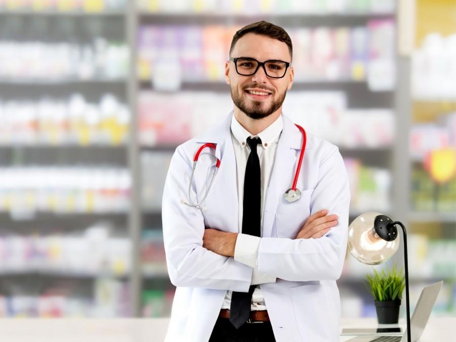 Ein Apotheker mit schwarzer Brille, freundlichem Blick und verschränkten Armen steht in seiner Apotheke. Neben ihm steht eine schwarze Schreibtischlampe und eine grüne Topfpfanze.