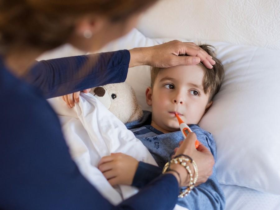 Ein kranker kleiner Junge liegt mit seinem Teddy im Bett und hat ein Fieberthermometer im Mund. Am Bett sitzt seine Mutter und prüft die Temperatur.