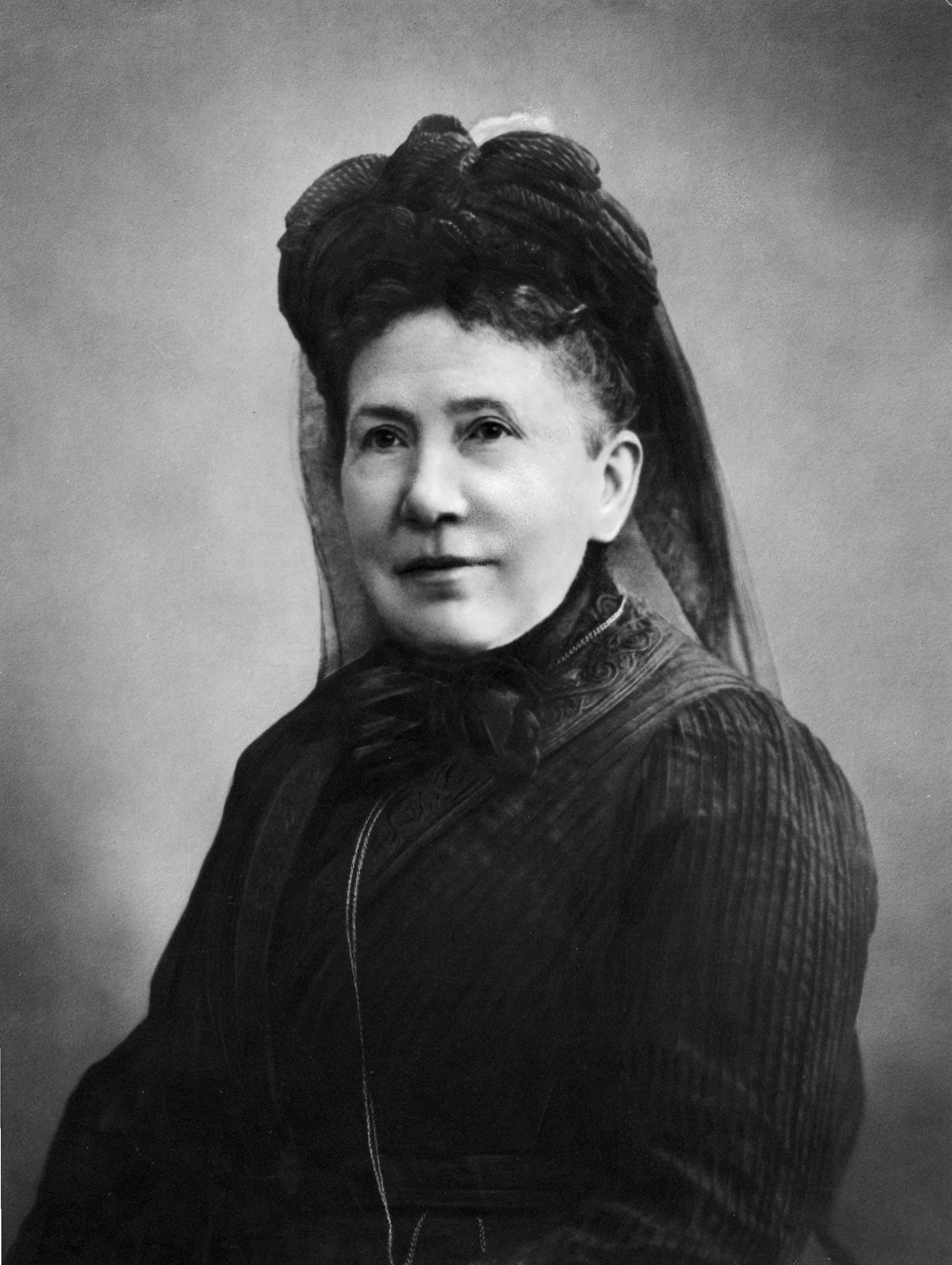 Auf dem Foto ist eine schwarz gekleidete Frau mittleren Alters zu sehen. Sie trägt ein hochgeschlossenes Oberteil und einen Damenhut mit Tüll.