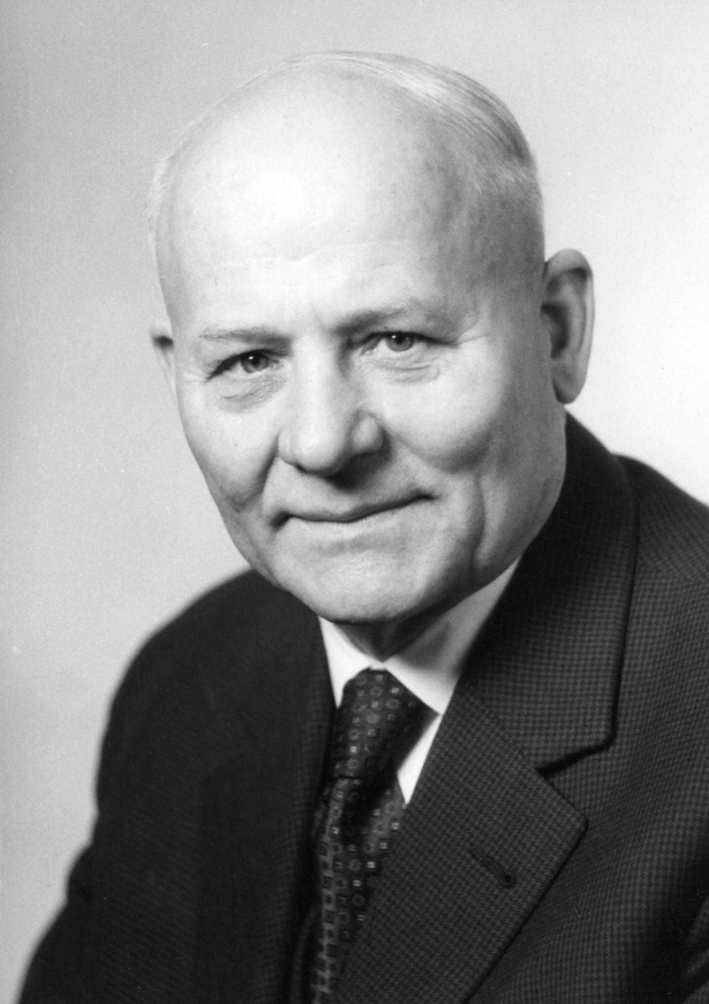Auf dem Portraitfoto ist ein älterer, lächelnder Herr ohne Haare zu sehen. Er trägt eine schwarze Anzugjacke, ein weißes Hemd und eine schwarze Krawatte.